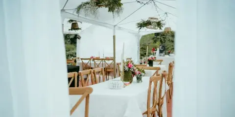 White event tent interior with draped fabric, wooden chairs, and tables set with white linens and floral centerpieces.