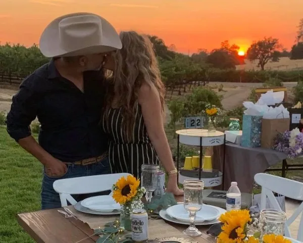 A couple standing at a decorated outdoor event table with white chairs and sunflowers during a sunset.