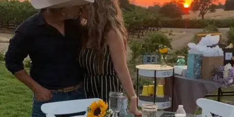 A couple standing at a decorated outdoor event table with white chairs and sunflowers during a sunset.