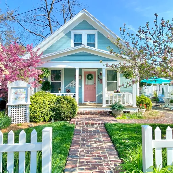 Exterior view of a blue house with a pink front door, white picket fence, and a brick walkway leading to the entrance.