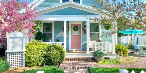 Exterior view of a blue house with a pink front door, white picket fence, and a brick walkway leading to the entrance.