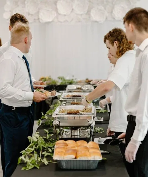 Catering staff serving guests at a buffet line with chafing dishes and bread rolls.