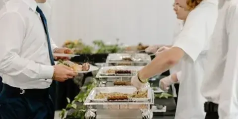 Catering staff serving guests at a buffet line with chafing dishes and bread rolls.