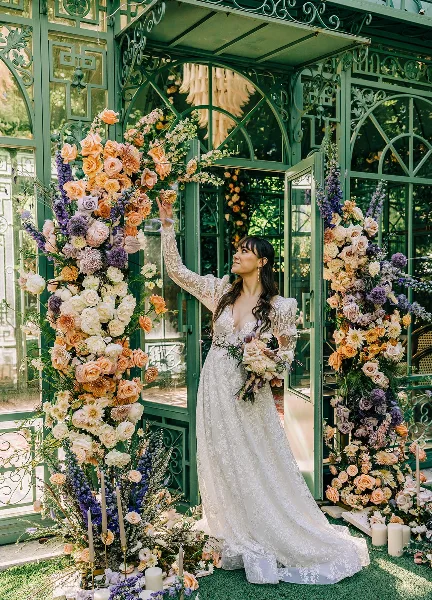 A bride in a white lace wedding dress stands in front of a green glass structure decorated with large floral arrangements of orange, white, and purple flowers.