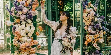 A bride in a white lace wedding dress stands in front of a green glass structure decorated with large floral arrangements of orange, white, and purple flowers.