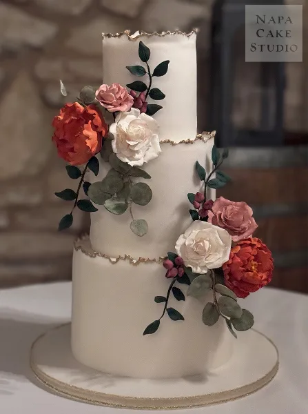 A three-tiered white wedding cake decorated with sculpted red and white flowers and gold leaf accents.