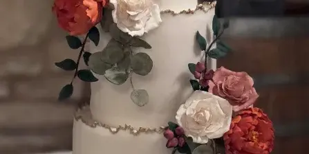 A three-tiered white wedding cake decorated with sculpted red and white flowers and gold leaf accents.