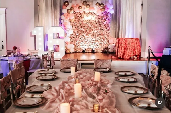 Interior view of an event venue showing a decorated stage with a floral wall and balloon arch, with a long dining table set with plates and candles in the foreground.