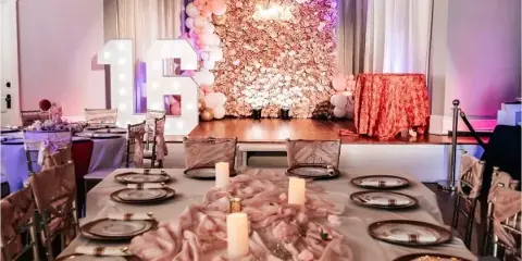 Interior view of an event venue showing a decorated stage with a floral wall and balloon arch, with a long dining table set with plates and candles in the foreground.