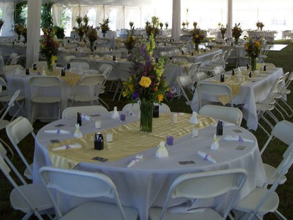 An outdoor event venue set up under a large white tent with multiple round tables covered in white and yellow linens, surrounded by white folding chairs and floral centerpieces.