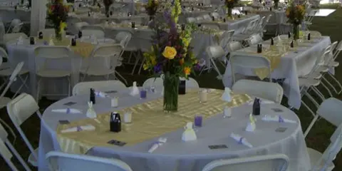 An outdoor event venue set up under a large white tent with multiple round tables covered in white and yellow linens, surrounded by white folding chairs and floral centerpieces.