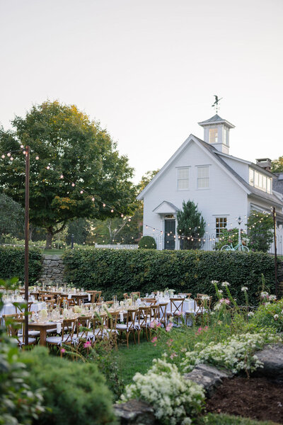 Outdoor wedding reception setup featuring string lights, floral centerpieces, and a white building in the background.