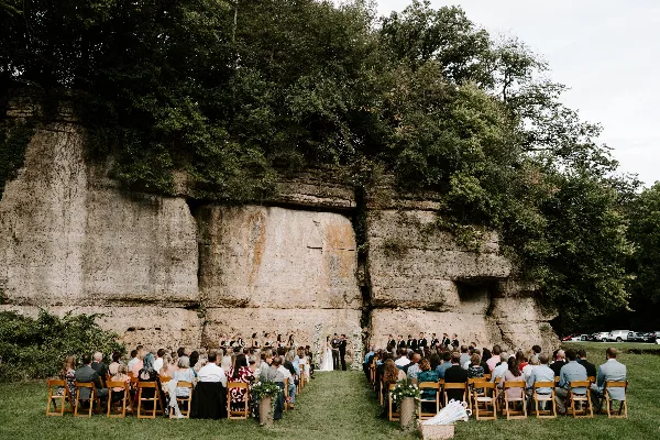 A wedding ceremony taking place outdoors in front of a large rock cliff face with guests seated in wooden chairs.