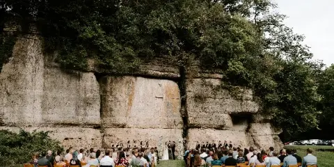 A wedding ceremony taking place outdoors in front of a large rock cliff face with guests seated in wooden chairs.