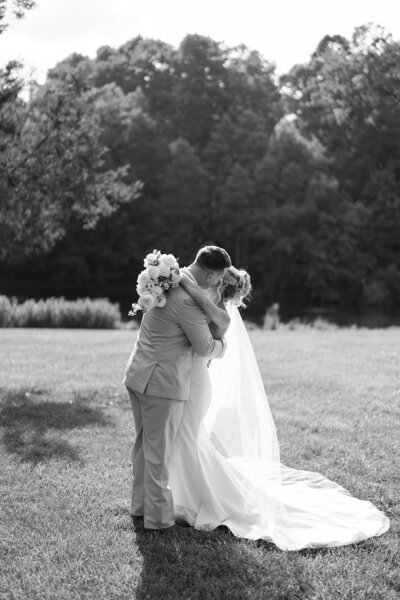 A black and white photograph of a bride and groom embracing in a grassy field with trees in the background.