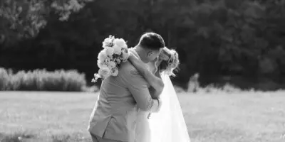 A black and white photograph of a bride and groom embracing in a grassy field with trees in the background.