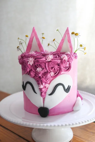 A pink custom cake decorated with a fox face, piped frosting flowers, and daisy toppers on a white cake stand.