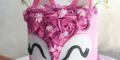 A pink custom cake decorated with a fox face, piped frosting flowers, and daisy toppers on a white cake stand.