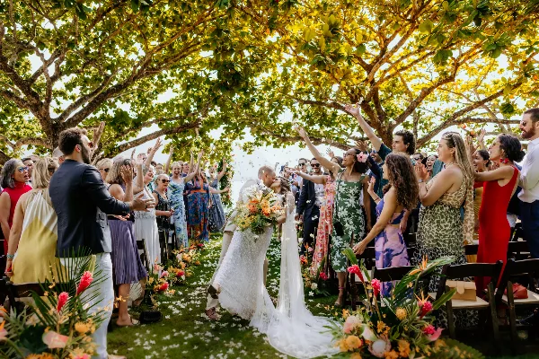 A bride and groom kiss during an outdoor wedding ceremony under a large tree while guests cheer and raise their hands.