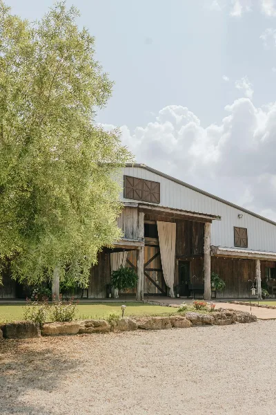 Exterior view of a rustic white and wood barn venue with a gravel driveway and landscaping.