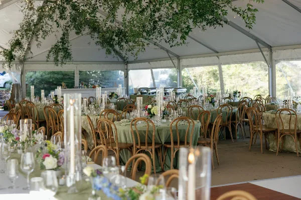 Interior view of a wedding reception tent with round tables, green linens, floral centerpieces, and tall candle holders.