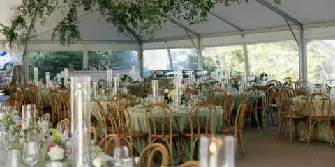 Interior view of a wedding reception tent with round tables, green linens, floral centerpieces, and tall candle holders.