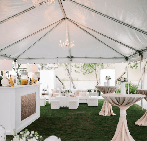 An outdoor wedding reception setup inside a white tent featuring a bar, chandeliers, and tables with draped linens.
