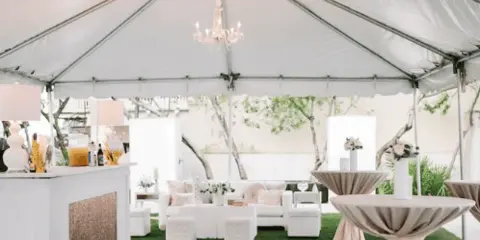 An outdoor wedding reception setup inside a white tent featuring a bar, chandeliers, and tables with draped linens.