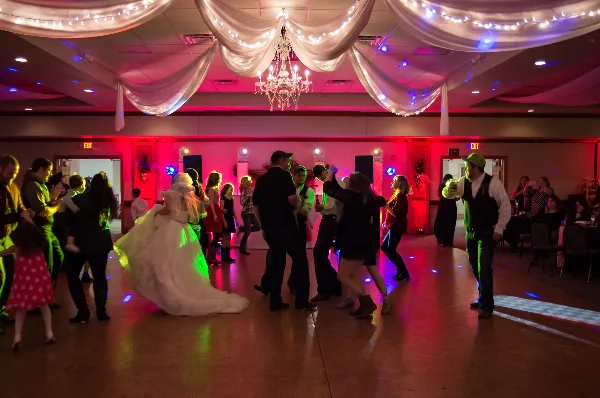 A wedding reception dance floor with guests dancing under colorful uplighting and a DJ booth in the background.