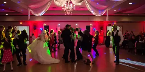 A wedding reception dance floor with guests dancing under colorful uplighting and a DJ booth in the background.