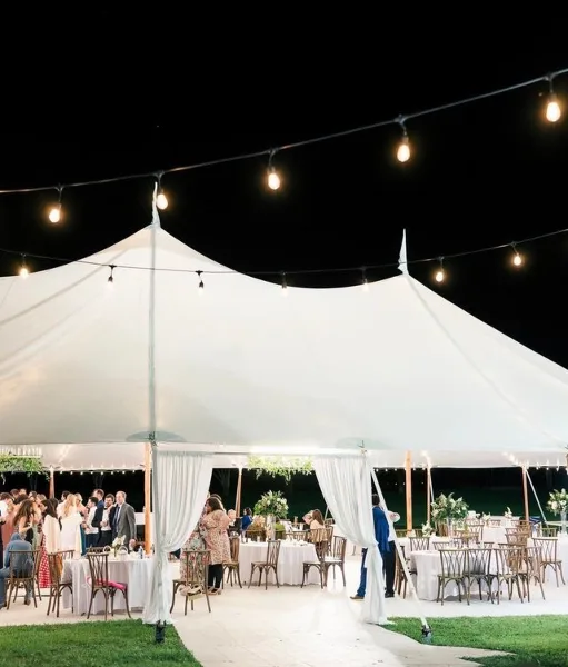 A large white event tent set up at night with string lights, containing tables and chairs for a wedding reception.