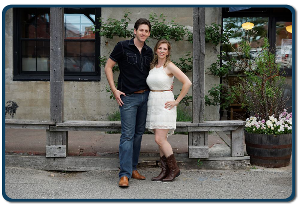 A man and a woman standing together in front of a rustic building, representing the Maine Music Makers band.