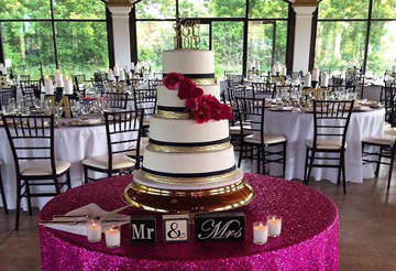 A tall white and gold wedding cake with red flowers displayed on a pink table in a banquet hall with set dining tables.