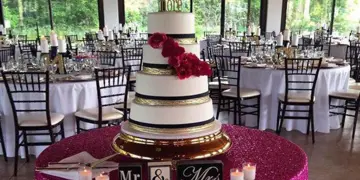 A tall white and gold wedding cake with red flowers displayed on a pink table in a banquet hall with set dining tables.