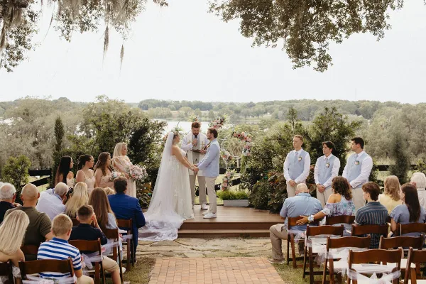 A bride and groom exchanging vows on a wooden stage with a floral arch, surrounded by seated guests and overlooking a lake.