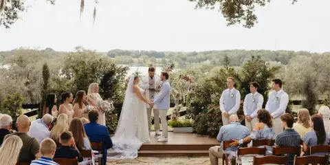 A bride and groom exchanging vows on a wooden stage with a floral arch, surrounded by seated guests and overlooking a lake.