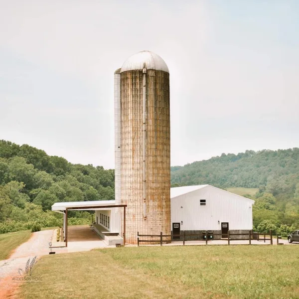 Exterior view of the Anderson Ridge event venue, showing a tall metal silo and a white barn building surrounded by green hills.