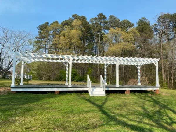 A white wooden pergola on a raised deck in a grassy outdoor area with trees in the background.