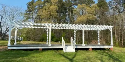 A white wooden pergola on a raised deck in a grassy outdoor area with trees in the background.