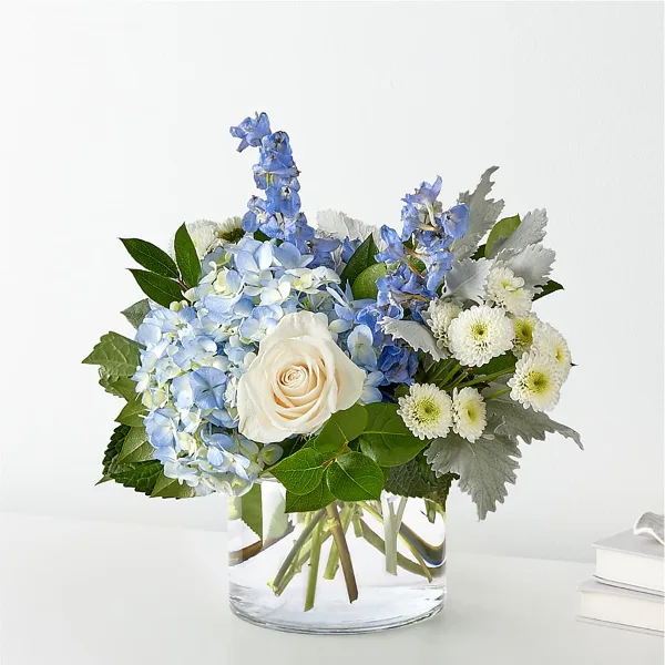 A floral arrangement featuring blue hydrangeas, a white rose, and delphiniums in a clear glass vase.
