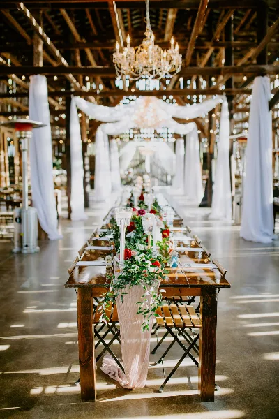 A long wooden banquet table decorated with a floral runner and candles, set in a rustic venue with white draping and chandeliers.