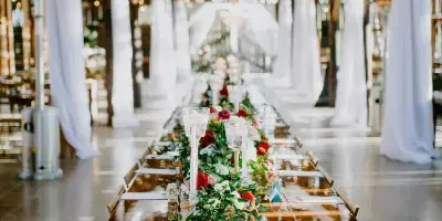 A long wooden banquet table decorated with a floral runner and candles, set in a rustic venue with white draping and chandeliers.