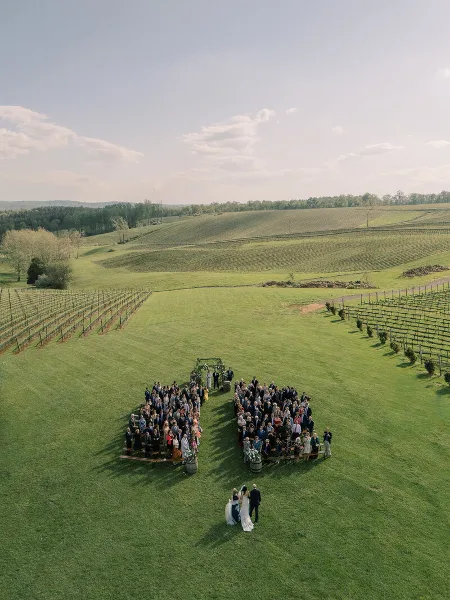 An aerial view of a wedding ceremony taking place in a vineyard, with the couple at the altar and guests seated on benches.