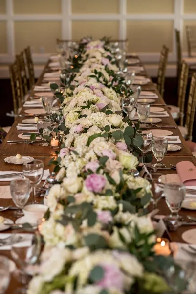 A long wooden dining table set for a wedding reception, featuring a lush floral runner with white and pink roses, eucalyptus, and candles.