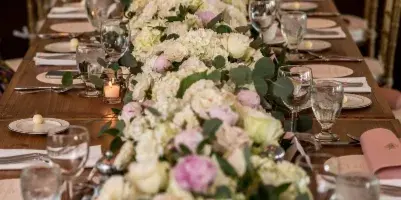 A long wooden dining table set for a wedding reception, featuring a lush floral runner with white and pink roses, eucalyptus, and candles.