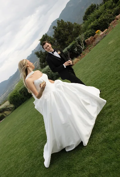 A bride in a white gown and a groom in a black tuxedo posing on a grassy hillside with mountains in the background.