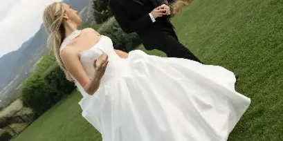 A bride in a white gown and a groom in a black tuxedo posing on a grassy hillside with mountains in the background.