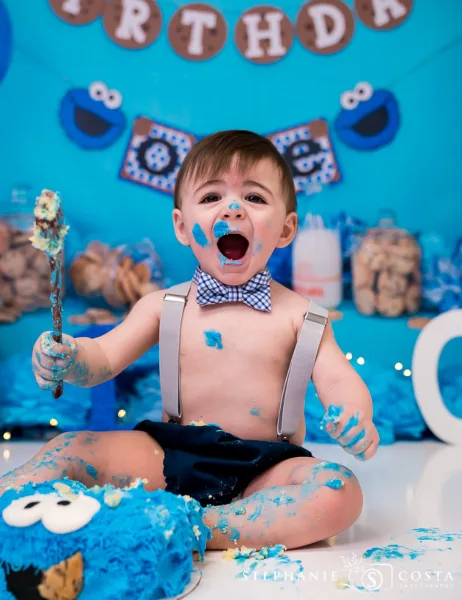 A toddler with blue frosting on their face and hands sits in front of a blue backdrop with a Cookie Monster themed cake smash setup.