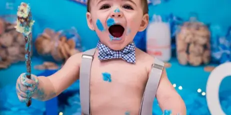 A toddler with blue frosting on their face and hands sits in front of a blue backdrop with a Cookie Monster themed cake smash setup.