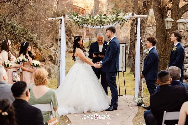 An officiant performing a wedding ceremony for a bride and groom under a floral arch in an outdoor garden setting.